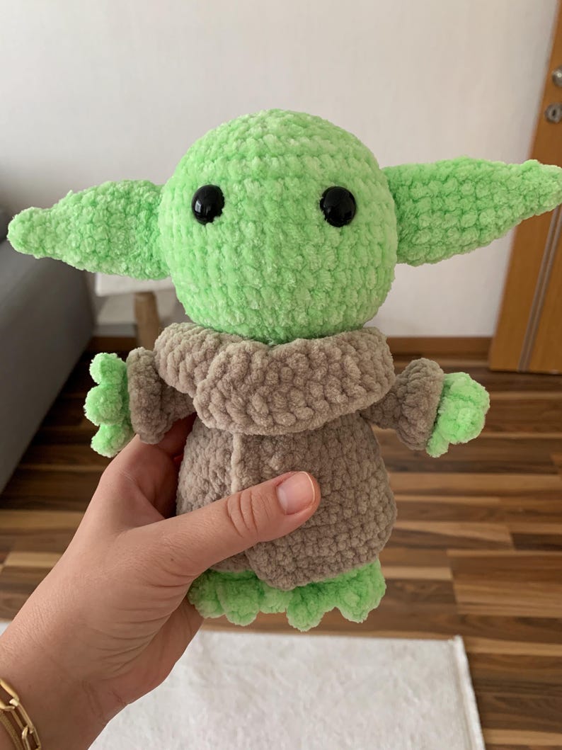 May include: A handmade green and brown crocheted doll, featuring large black eyes and oversized ears. The doll is wearing a brown coat and has green hands and feet. The doll is a character from a popular science fiction franchise.