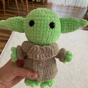 May include: A handmade crochet doll in the likeness of a popular character. The doll features a light green head and ears, black eyes, and a tan-colored outfit. The doll is approximately 8 inches tall.