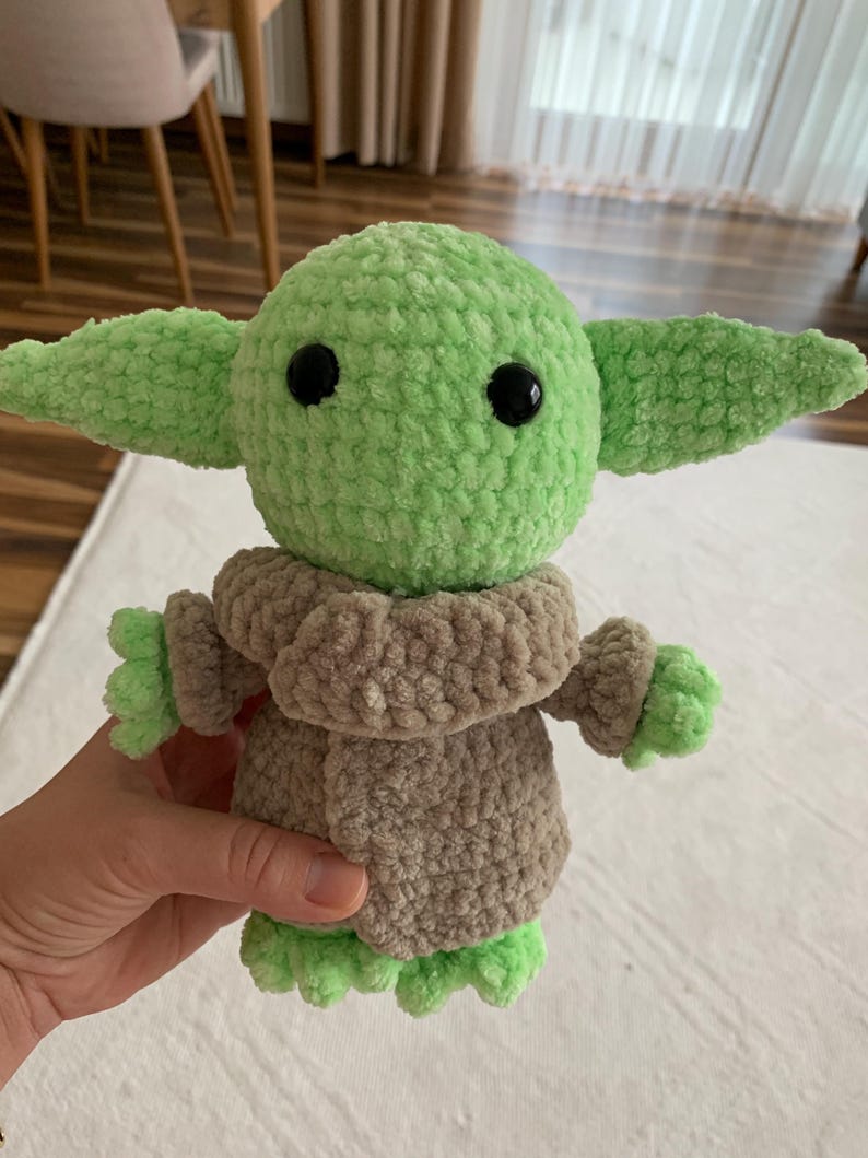 May include: A handmade crochet doll in the likeness of a popular character. The doll features a light green head and ears, black eyes, and a tan-colored outfit. The doll is approximately 8 inches tall.