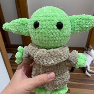 May include: A handmade green and tan crocheted doll with large ears and black eyes. The doll is wearing a tan robe and has green hands and feet. The doll is a character from a popular science fiction franchise.