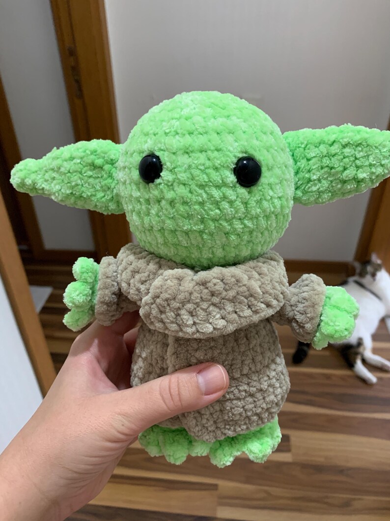 May include: A handmade green and tan crocheted doll with large ears and black eyes. The doll is wearing a tan robe and has green hands and feet. The doll is a character from a popular science fiction franchise.