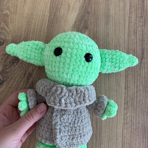 May include: A crocheted plush toy of a character with large ears, green skin, and black eyes. The character wears a gray coat. The toy is held by a hand against a wooden background. The toy is approximately 6 inches tall.