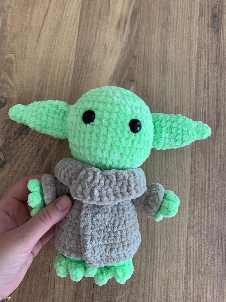 May include: A crocheted plush toy of a character with large ears, green skin, and black eyes. The character wears a gray coat. The toy is held by a hand against a wooden background. The toy is approximately 6 inches tall.
