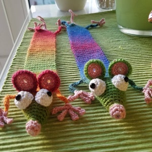 May include: Two crocheted mouse bookmarks with rainbow colored tails. The mice have green bodies, pink noses, white eyes, and black pupils. The bookmarks are on a green textured surface.