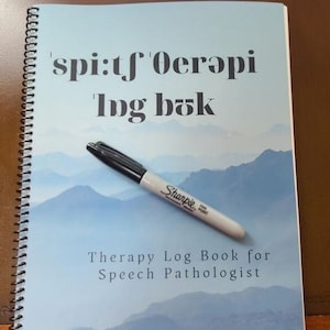 May include: A blue and white spiral bound notebook with the text "'spi:t'f'oerapi 'lpg bøk" and "Therapy Log Book for Speech Pathologist" on the cover. A black and white marker is lying on top of the notebook.