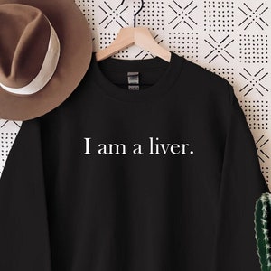 May include: Black sweatshirt with the text "I am a liver." displayed in white. A brown hat with a white band hangs on the left. A cactus in a white pot is on the right. The background is a white and black patterned wall.