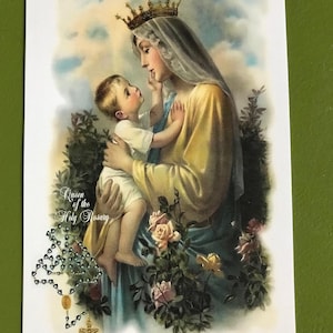 May include: A religious print depicting a woman wearing a crown and veil, holding a child. The woman is dressed in a yellow and blue robe, with a halo. The image includes roses, greenery, and a rosary. Text reads "Queen of the Holy Rosary."