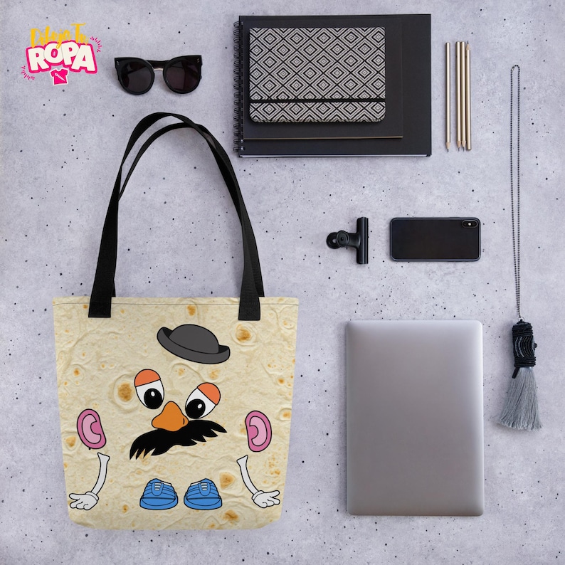 May include: A beige tote bag featuring a cartoon character design. The character has a black mustache, a gray bowler hat, and blue shoes. The bag has black handles and is likely made of fabric.