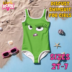 Peut inclure: Un maillot de bain vert avec un visage de dessin animé de dégoût, une bordure blanche et le texte "DISGUST SWIMSUIT FOR GIRLS" et "SIZES 2T-7". Des lunettes de soleil roses et des coquillages sont sur le sable.