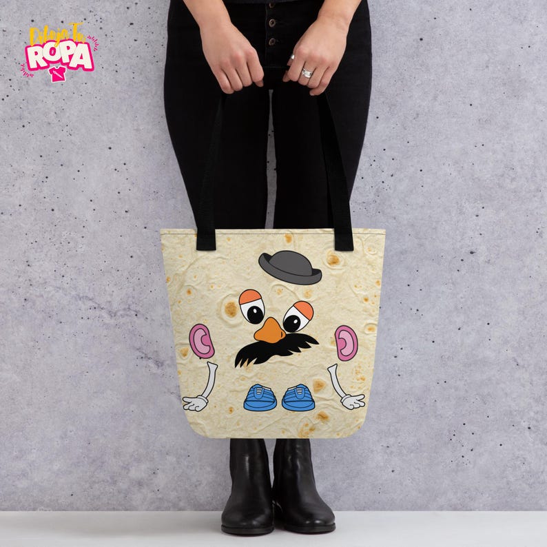 May include: A beige tote bag featuring a cartoon Mr. Potato Head design. The design includes a black mustache, a gray bowler hat, and blue shoes. The bag has black handles and the text "ROPA" in the upper left corner.