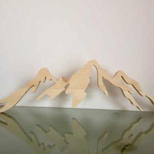 以下が含まれることがあります： 自然な木から切り出された、薄い茶色の木製の山脈のシルエット。山脈は反射面の上に置かれています。