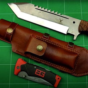 May include: A large hunting knife with a serrated blade and wooden handle rests above a brown leather sheath. A smaller folding knife with a black and red handle is below. The knives are on a green surface.