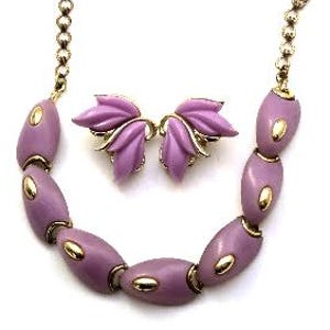 Könnte beinhalten: Ein Vintage-Schmuckset mit Halskette und passenden Ohrringen. Die Halskette hat lavendelfarbene ovale Perlen mit goldfarbenen Akzenten, verbunden durch eine goldfarbene Kette. Die Ohrringe sind wie stilisierte Lavendelblüten geformt. Das Set wird auf weißem Hintergrund präsentiert.