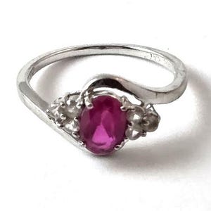 May include: A silver ring with a large oval, bright pink gemstone as the focal point. The ring has a swirling design with small accent stones on either side of the main stone. The ring is set against a white background.