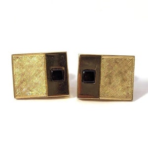 May include: A pair of gold-coloured cufflinks. Each cufflink has a textured gold section and a dark brown section with a small, square black stone. The cufflinks are rectangular and set against a white background.