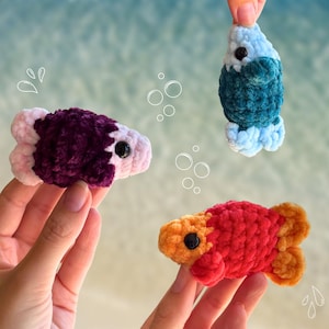 Könnte beinhalten: Drei handgefertigte Häkelfische in leuchtenden Farben: Lila und Rosa, Türkis und Hellblau sowie Rot und Orange. Jeder Fisch hat ein schwarzes Auge und ein strukturiertes, plüschiges Aussehen. Die Fische werden vor einem verschwommenen, hellblauen Hintergrund gehalten.