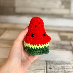 Könnte beinhalten: Ein handgefertigtes Plüsch-Wassermelonenstück. Das Spielzeug hat eine rote Oberseite, eine grüne und gelbe Schale und schwarze Kerndetails. Das weiche, strukturierte Spielzeug wird in einer Hand gehalten.