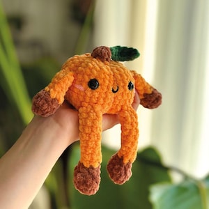 Könnte beinhalten: Ein handgefertigtes orangefarbenes Häkeltier mit einem lächelnden Gesicht, schwarzen Augen und braunen Füßen und Händen. Das Spielzeug hat einen kleinen braunen Stiel und ein grünes Blatt oben. Das Plüschtier wird in einer Hand gehalten.