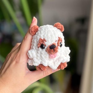 Könnte beinhalten: Ein handgefertigtes weiß-braunes Plüschtier in Form eines Hundes. Das Spielzeug hat schwarze Augen, eine rosa Zunge und braune Akzente an Ohren und Gesicht. Das Plüschtier wird in einer Hand gehalten.