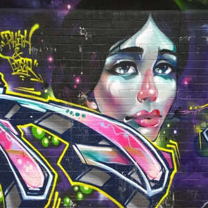 May include: A colourful graffiti mural on a brick wall features a woman's face with blue eyes and pink lips. The mural also includes abstract shapes in pink, blue, yellow, and green.