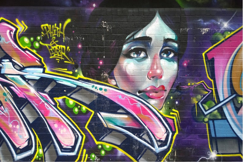 May include: A colourful graffiti mural on a brick wall features a woman's face with blue eyes and pink lips. The mural also includes abstract shapes in pink, blue, yellow, and green.