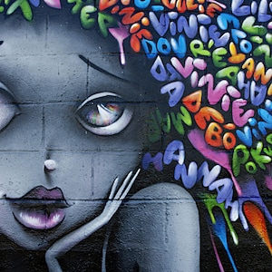 May include: A colourful graffiti mural of a woman's face with large eyes and a pink mouth. The background is a brick wall with a colourful spray paint design of letters and words.