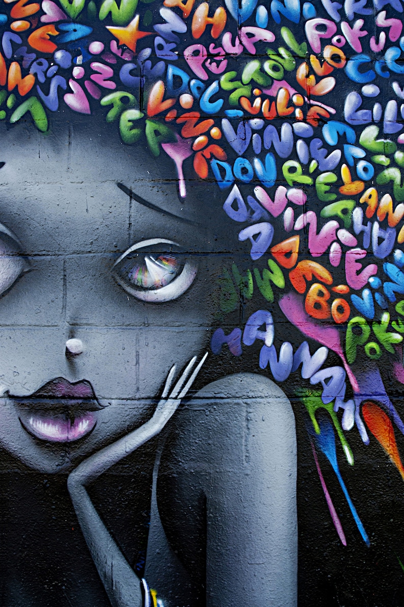 May include: A colourful graffiti mural of a woman's face with large eyes and a pink mouth. The background is a brick wall with a colourful spray paint design of letters and words.