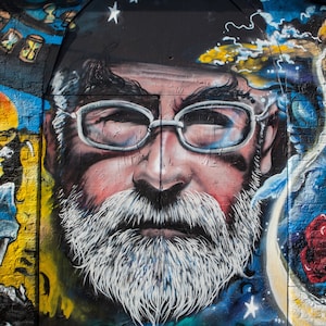 May include: A colourful street art mural featuring a close-up of a man's face with a white beard and glasses. The mural is painted on a brick wall and includes other details like stars, moons, and a figure with a skeleton hand.