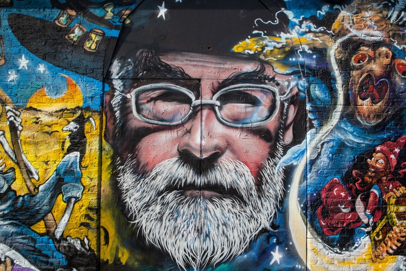 May include: A colourful street art mural featuring a close-up of a man's face with a white beard and glasses. The mural is painted on a brick wall and includes other details like stars, moons, and a figure with a skeleton hand.