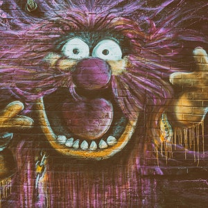 May include: A colourful graffiti mural on a brick wall depicts a cartoon character with a large, open mouth, a pink nose, and wild purple hair. The character is painted in a vibrant style with dripping paint effects.