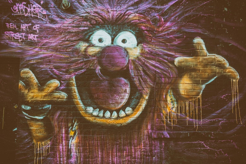 May include: A colourful graffiti mural on a brick wall depicts a cartoon character with a large, open mouth, a pink nose, and wild purple hair. The character is painted in a vibrant style with dripping paint effects.