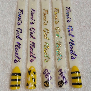 May include: Four white sticks with the text "Toni's Gel Nails" printed on them. Each stick has a different design on a yellow nail: black and yellow stripes, white hearts with black outlines, a bee with the word "Bee" above it, and a bee with the word "Mine" above it.