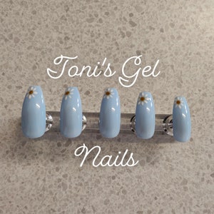 May include: Set of six light blue gel nails with a white daisy design. The nails are displayed on a clear stand. The text "Toni's Gel Nails" is written above the nails.