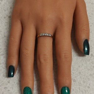 May include: A hand with green glitter nail polish on four fingers and gold glitter nail polish on one finger. A thin gold band with small clear stones is worn on the ring finger.