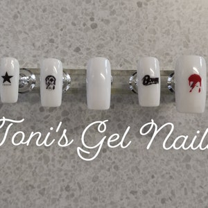 May include: Five white fake nails with different designs. The nails are arranged in a row on a silver display stand. The designs include a star, a dog, the word "BOWIE", and a red and black lightning bolt. The text "Toni's Gel Nails" is written below the nails.
