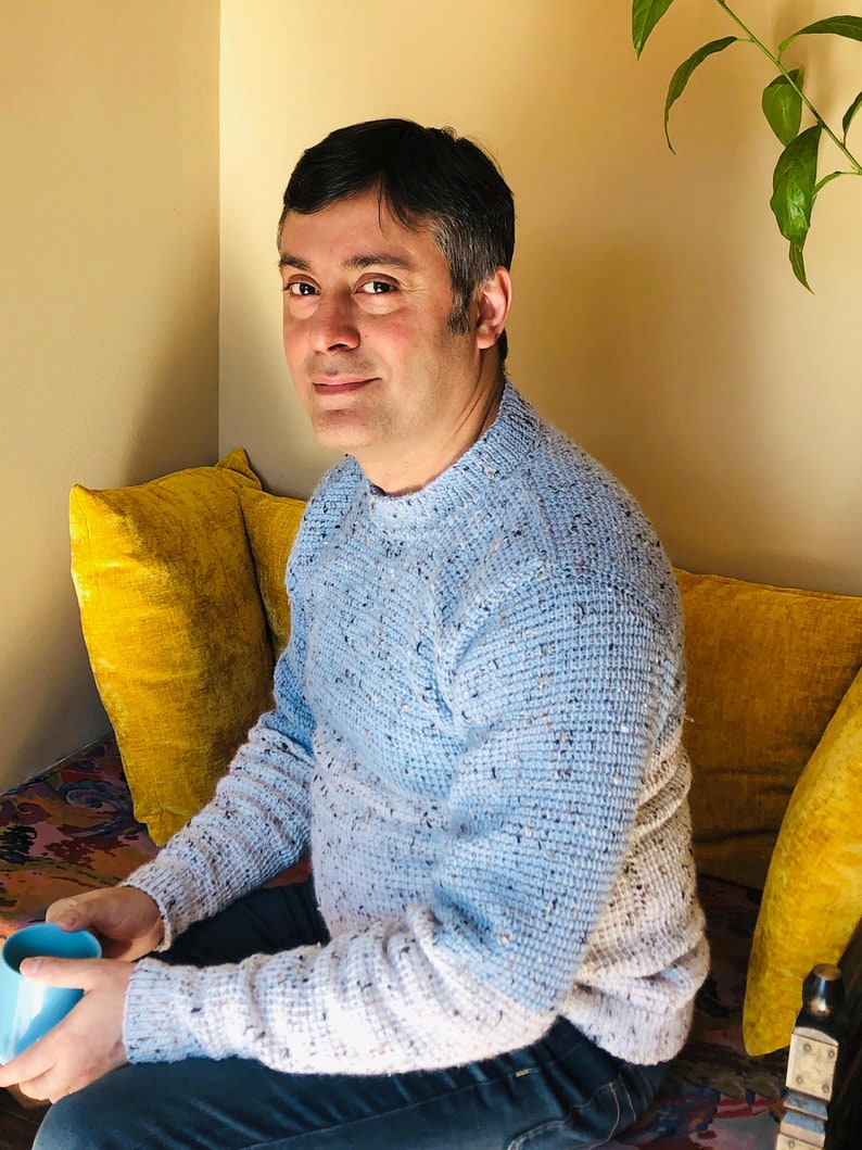 May include: A man wearing a blue and white speckled sweater sits on a yellow couch. He is holding a light blue mug in his left hand.