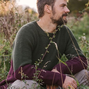May include: A man wearing a green and burgundy sweater sits in a field of tall grass. The sweater has a waffle knit texture and the sleeves are a darker burgundy color.