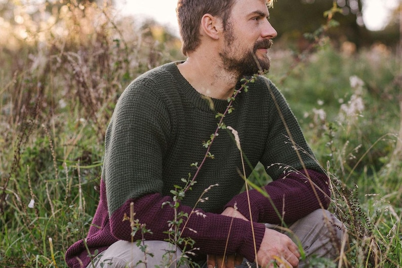May include: A man wearing a green and burgundy sweater sits in a field of tall grass. The sweater has a waffle knit texture and the sleeves are a darker burgundy color.