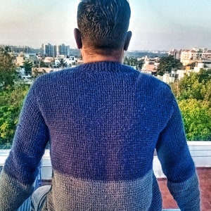 May include: A person wearing a blue and gray sweater, sitting on a concrete surface and looking out at a cityscape.