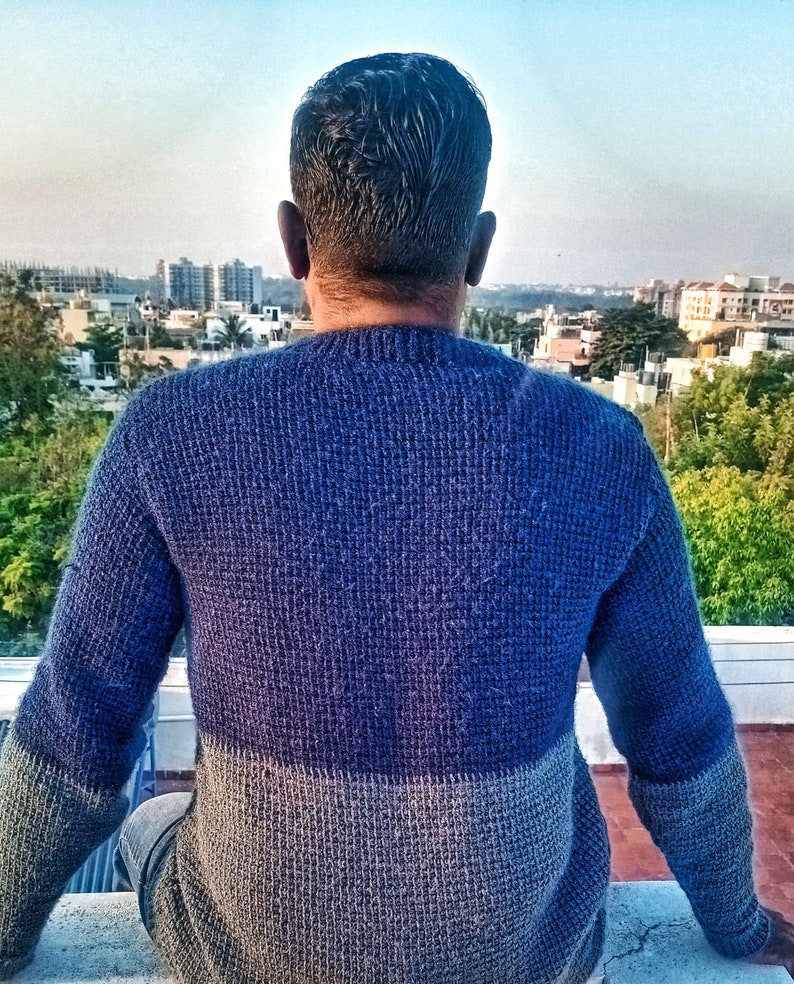 May include: A person wearing a blue and gray sweater, sitting on a concrete surface and looking out at a cityscape.