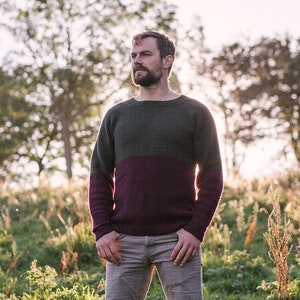 May include: A man wearing a green and burgundy sweater stands in a field of tall grass. He is looking off to the side, and the sun is shining behind him.