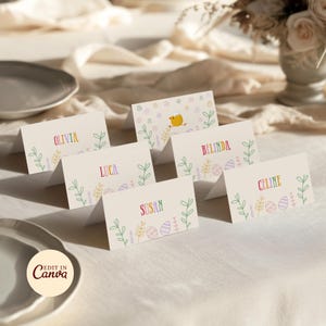 May include: Cream-colored Easter place cards with names in rainbow colors. Each card features a floral and egg design. One card has a yellow chick illustration. The cards are arranged on a table with plates and flowers.