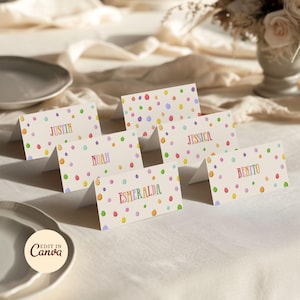 May include: Six folded place cards with names in rainbow colors, including Justin, Noah, Esmeralda, Jessica, and Benito. Each card has a cream background with colorful polka dots and diagonal lines. The cards are arranged on a white tablecloth.