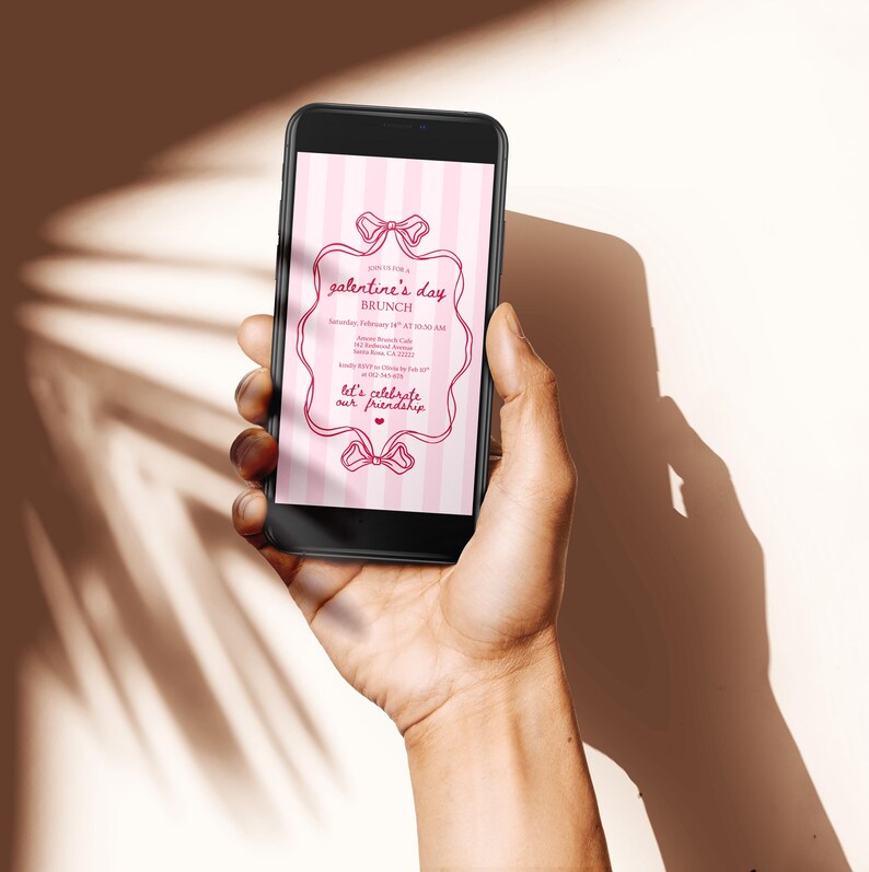 May include: A black smartphone displays a pink and white striped invitation for a Galentine's Day brunch. The invitation features a decorative frame with bows and text that reads "Let's celebrate our friendship."