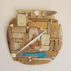 Könnte beinhalten: Eine runde Wanduhr aus Treibholz und Muscheln. Das Zifferblatt der Uhr ist ein blauer Fisch mit einem weißen Zeiger.