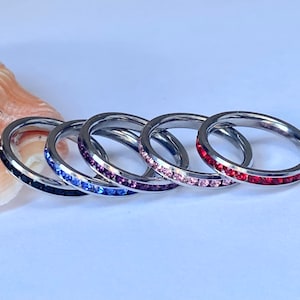 May include: Four stainless steel rings with a single row of colourful crystals. The rings are stacked on top of each other, with the red ring on top, followed by the pink, purple, and black rings.