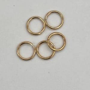 May include: Close-up of several gold-coloured jump rings, some open and some closed. The rings are arranged on a white surface. These small, circular components are commonly used in jewellery making and crafting.