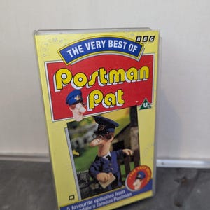 Könnte beinhalten: Eine VHS-Kassette mit "The Very Best of Postman Pat". Das gelbe Gehäuse hat roten und blauen Text mit Bildern der Zeichentrickfigur. Das Band enthält 5 Lieblingsfolgen von Greendales berühmtem Postboten.