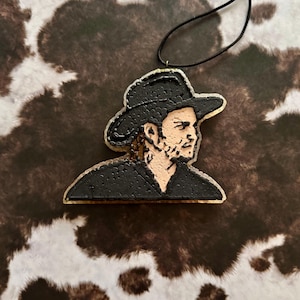 May include: Black and white beaded car air freshener featuring a portrait of a man wearing a cowboy hat.