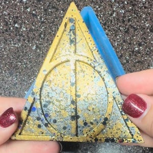 May include: A gold glitter resin casting of the Deathly Hallows symbol from the Harry Potter series. The casting is in a triangle shape with a circle inside and a vertical line through the circle. The casting is being removed from a blue silicone mold.